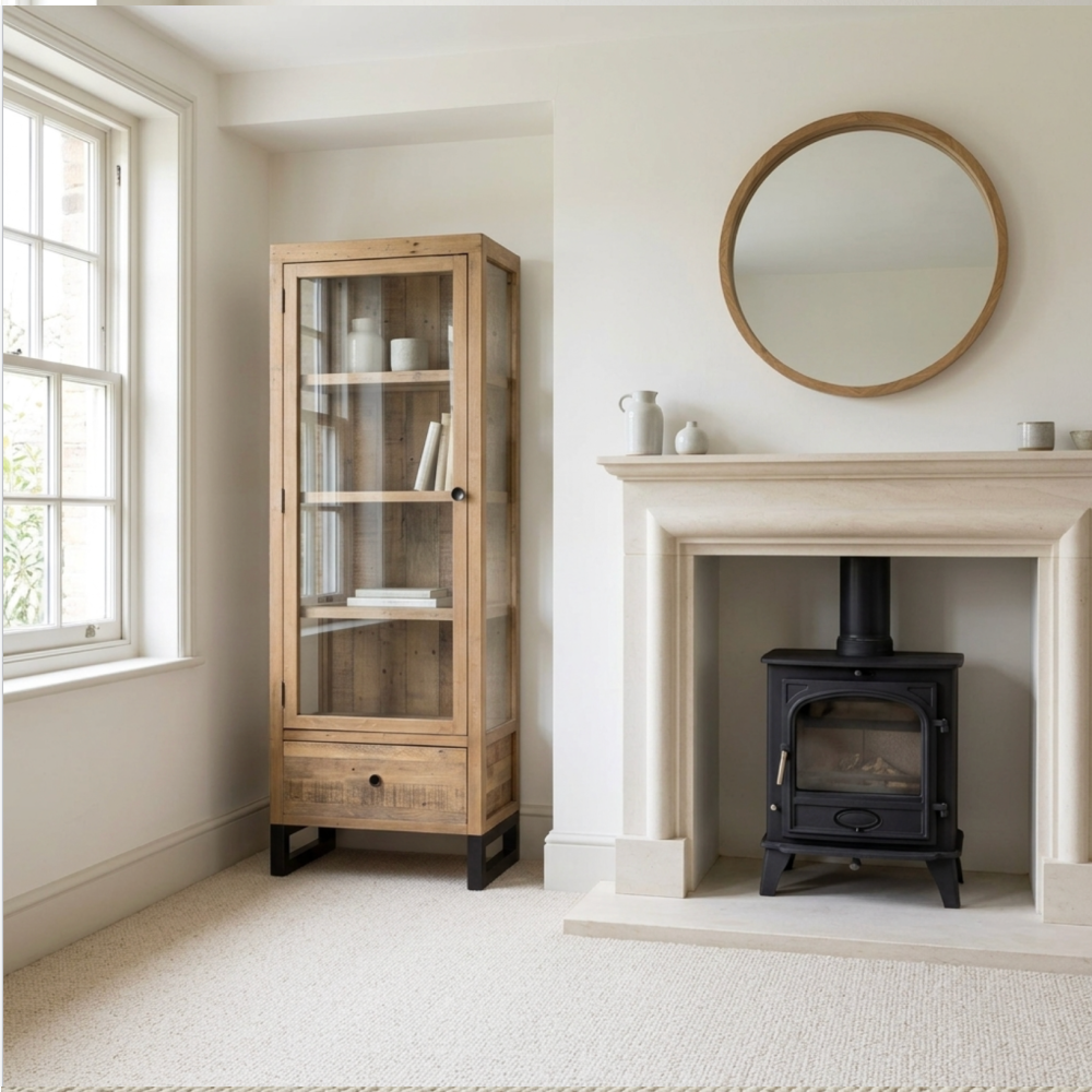 Wooden display cabinet next to a fireplace with a round mirror above it in a room with a window.
